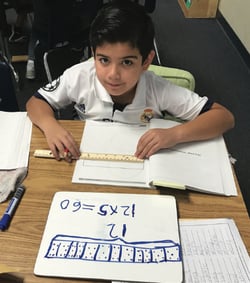 A student sitting at a table holding a ruler on a workbook page with a whiteboard with a math problem on it in front of the workbook.