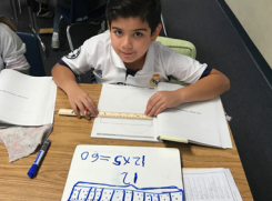 Student sitting at table with ruler, workbook, and whiteboard with math problem on table.