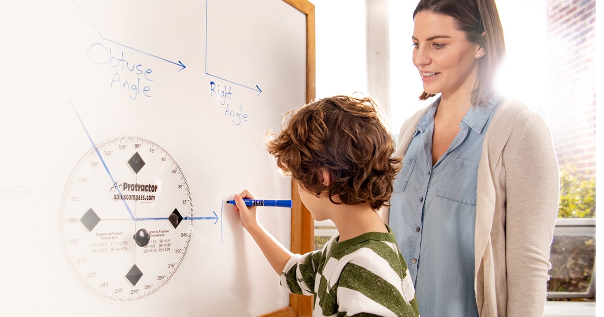 A teacher and student standing at a whiteboard. The students is holding a marker and writing on the whiteboard. There is a protractor on the whiteboard to the left of where he is writing. 