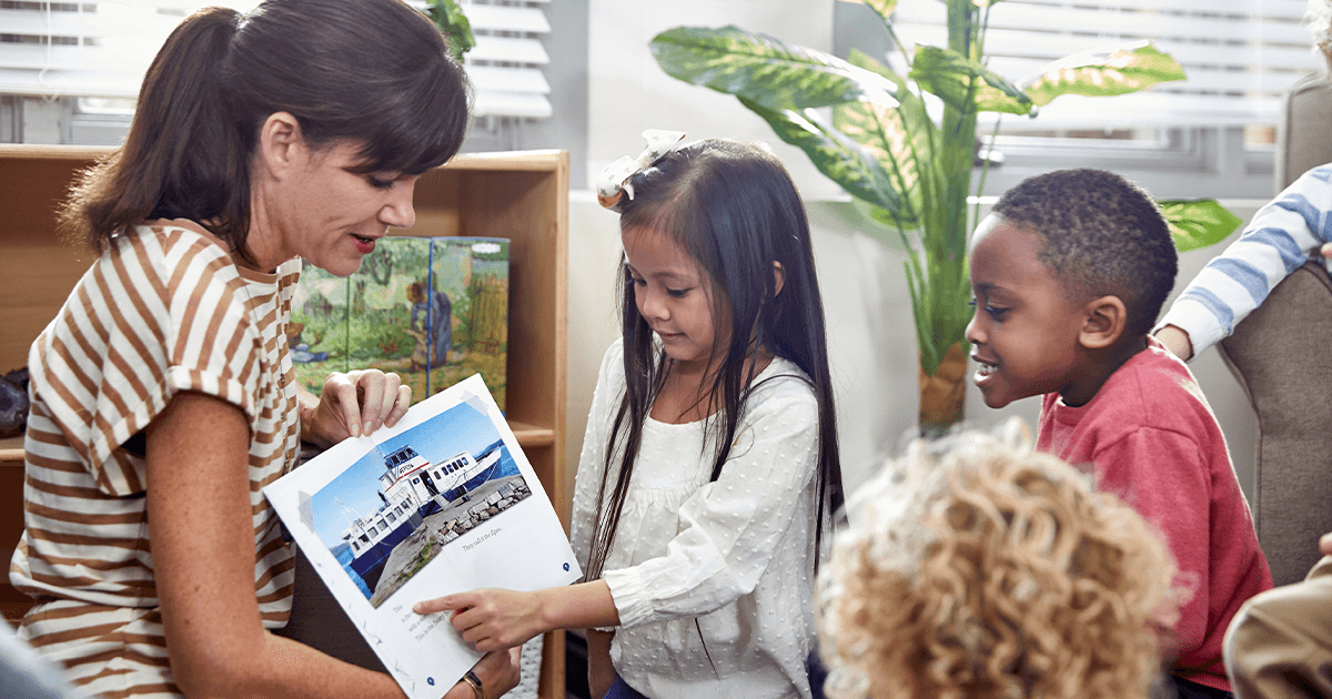 Teacher holding a book out to a group of students. A young girl is pointing at one of the pages of the book.