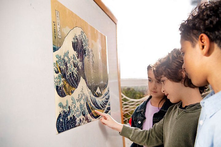 Three students examining a framed print of a large ocean wave on a wall, with one child pointing at details in the artwork.