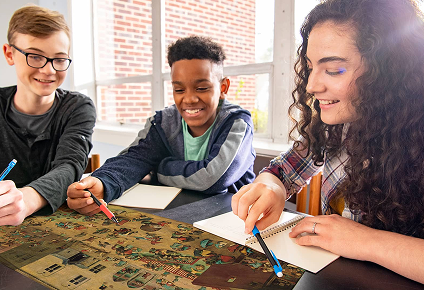 Three students smiling and working together at a table, pointing with colored pencils at a detailed illustrated map while taking notes in a bright classroom.