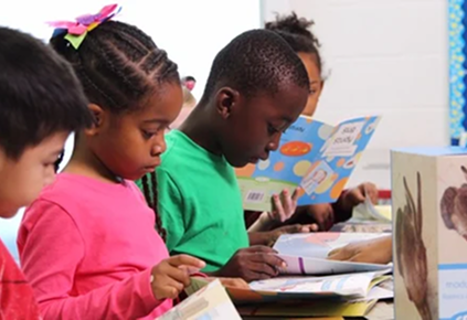 Four elementary school students sitting side by side at a desk, quietly reading colorful books in a classroom.