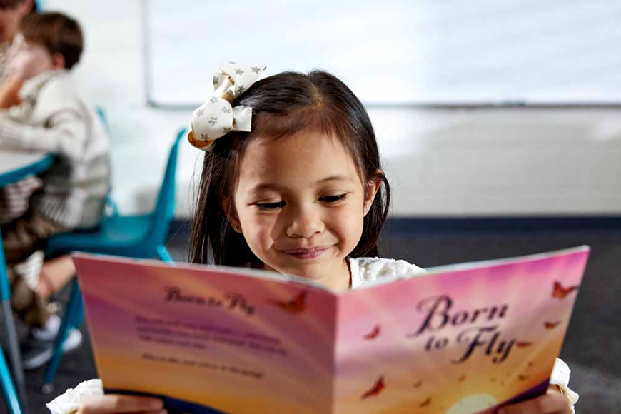 Young girl with a white bow in her hair smiling while reading a pink book titled “Born to Fly” in a classroom, with other children blurred in the background. 