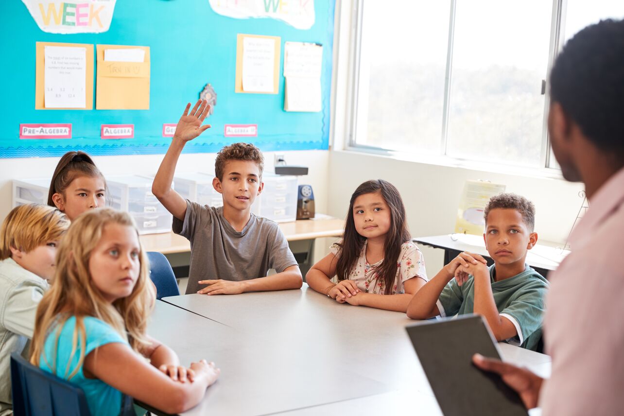 A group of six students sitting at a table while the teacher engages with them. The student in the middle raises his hand. The teacher holds a tablet in their hand.