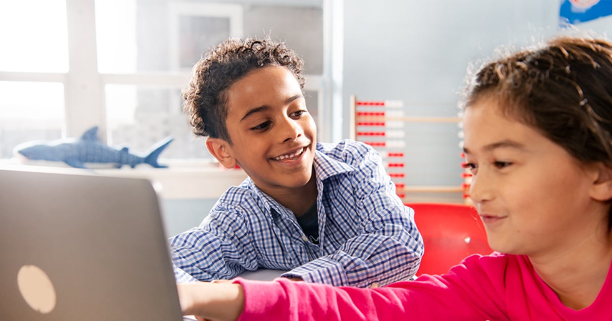 A boy and girl sitting at a desk looking at a shared computer screen. 